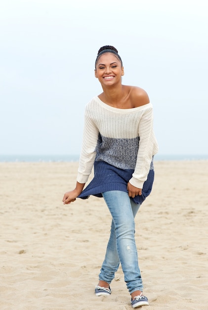 Feliz mujer joven caminando en la playa en jeans y suéter