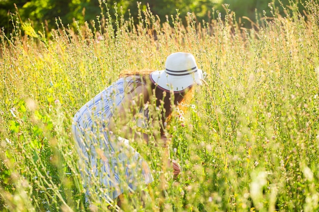 Feliz mujer joven con pelo largo con sombrero y vestido tira de sus manos hacia las plantas mientras camina