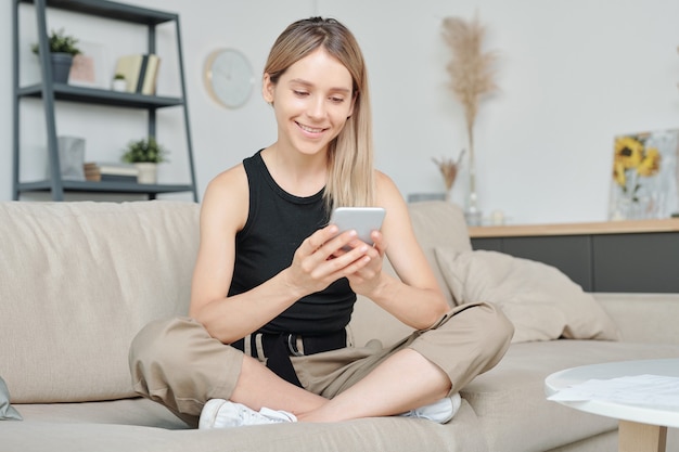 Feliz mujer joven en ropa casual sentada en el sofá con las piernas cruzadas y mirando a través de fotos en el teléfono inteligente mientras se queda en casa