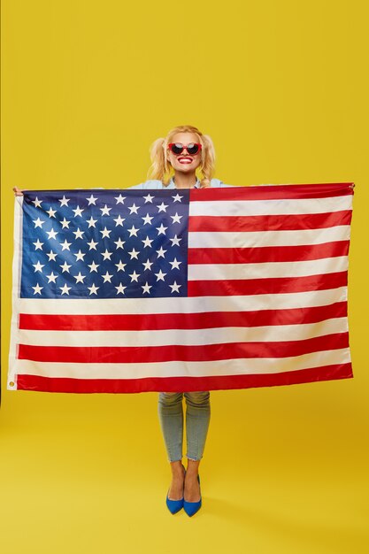 Feliz mujer joven en ropa de mezclilla sosteniendo la bandera de Estados Unidos aislado sobre fondo amarillo