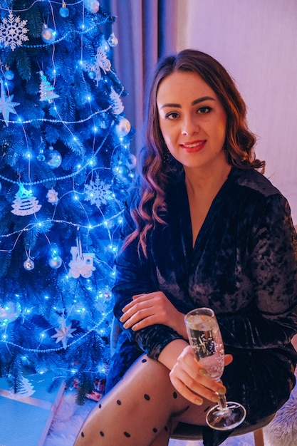 Feliz mujer joven sentada con copa de champán y luz de Bengala junto al árbol de Navidad. Celebración de la fiesta de año nuevo. Vacaciones de invierno de Navidad. Vispera de Año Nuevo.