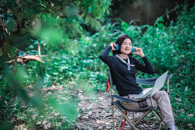 Feliz mujer joven sentada en una silla de camping usando auriculares escuchando música de una computadora portátil mientras se relaja acampando en el bosque