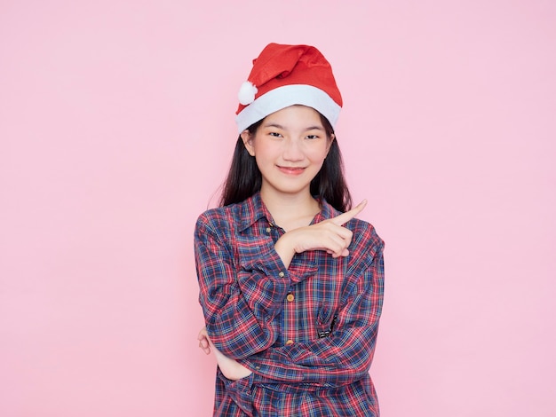 Feliz mujer joven con sombrero de santa mirando a la cámara sosteniendo el dedo apunta al lugar vacío para el texto sobre fondo rosa.