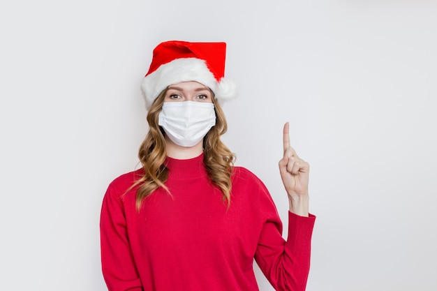 Feliz mujer joven con sombrero de santa, suéter rojo con máscara protectora médica señala con el dedo a la copia del texto aislado sobre fondo blanco de estudio. Concepto de cuarentena de coronavirus