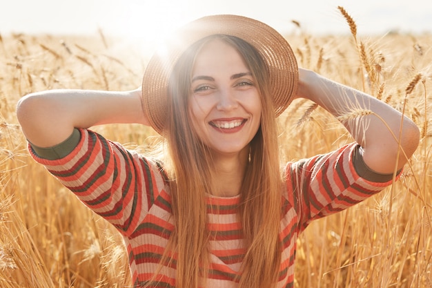 Feliz mujer joven en traje de rayas y sombrero para el sol disfrutando del sol en el campo de trigo, mantiene sus manos sobre la cabeza