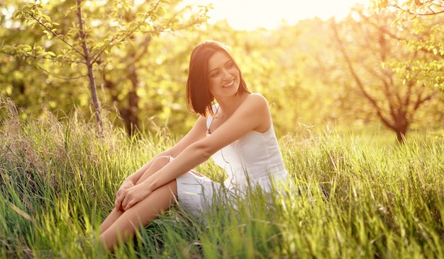 Feliz mujer joven en vestido blanco sonriendo y mirando a otro lado mientras está sentado sobre la hierba verde fresca en un día soleado de verano en el campo y descansando