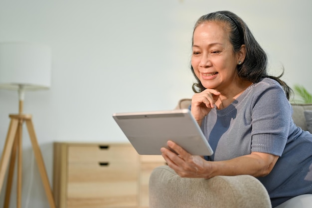 Feliz mujer jubilada asiática viendo películas en su tableta digital en el sofá