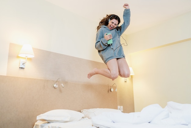 Feliz mujer latina usa auriculares saltando en la cama escuchando música cantando canciones con los ojos cerrados