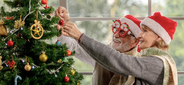 Feliz mujer madura y anciano con gorro de Papá Noel sonriendo y colgando adornos en el árbol de coníferas mientras se prepara para la celebración de Navidad en casa