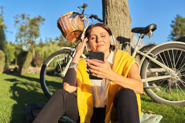 Feliz mujer madura con auriculares de ojos cerrados sentado en el parque, hermosa mujer cantando y escuchando música. Felicidad, relajación, ocio, estilo de vida, personas de mediana edad