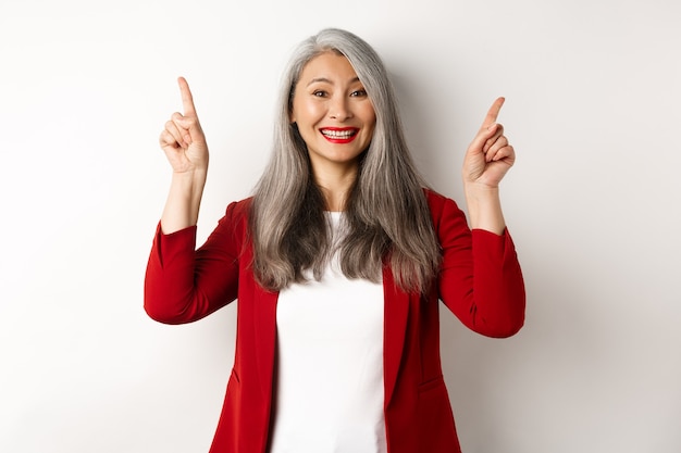 Feliz mujer madura en chaqueta roja y maquillaje, sonriendo y mostrando publicidad en la parte superior, apuntando con el dedo hacia el logo, fondo blanco.