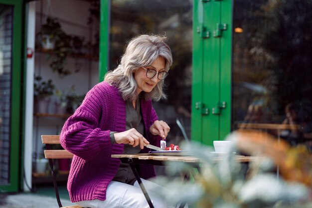 Feliz mujer madura con gafas come un delicioso postre en la terraza de un café al aire libre