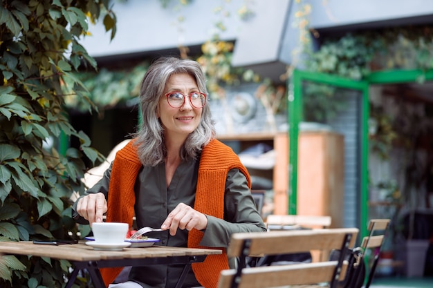 Feliz mujer madura con gafas come postre sentado en una mesa pequeña al aire libre