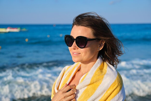 Feliz mujer madura relajándose en la playa, mujer sonriente en gafas de sol con toalla sobre los hombros, copie el espacio. Fondo de paisaje marino al atardecer