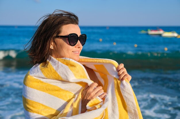 Feliz mujer madura relajándose en la playa, mujer sonriente en gafas de sol con toalla sobre los hombros, copie el espacio. Fondo de paisaje marino al atardecer