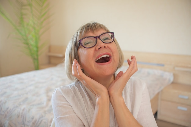 Feliz mujer mayor con cabello gris relajante sonriendo está mirando a la cámara en su casa en el dormitorio.