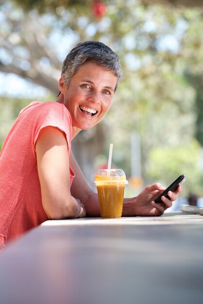 Feliz mujer mayor sentada afuera con bebida y usando un teléfono móvil