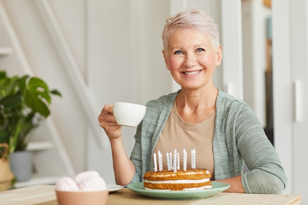 Feliz mujer mayor sonriendo a la cámara mientras está sentado en la mesa y bebiendo té con pastel