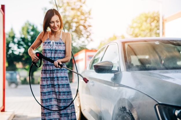 Feliz mujer de mediana edad lavando el coche en la estación de lavado de coches con máquina de agua a alta presión.