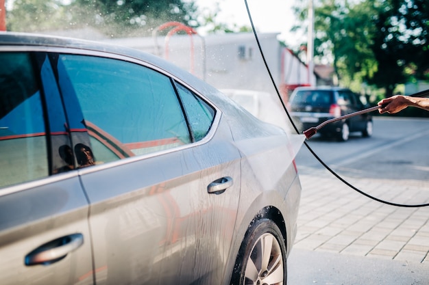 Feliz mujer de mediana edad lavando el coche en la estación de lavado de coches con máquina de agua a alta presión.