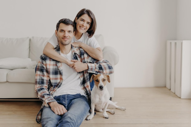 Feliz mujer morena abraza a su esposo con amor estando de buen humor sonríe positivamente El esposo, la esposa y el perro posan juntos en la sala de estar de la nueva vivienda disfrutan de la comodidad Pareja enamorada interior