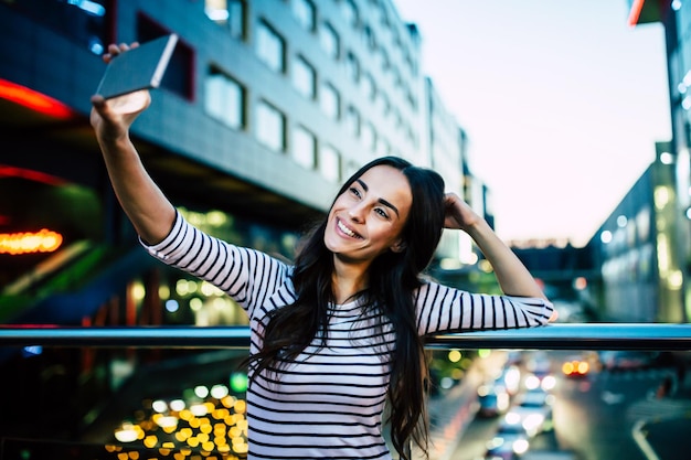 Feliz mujer morena atractiva sonriente hace selfie en su teléfono inteligente en el fondo de la ciudad de noche