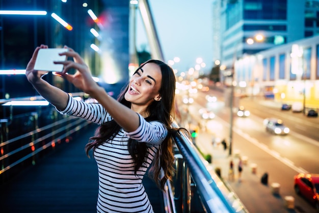 Feliz mujer morena atractiva sonriente hace selfie en su teléfono inteligente en el fondo de la ciudad de noche