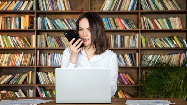 Feliz mujer morena en elegante blusa blanca habla por teléfono móvil cerca de la computadora portátil contemporánea en la mesa contra grandes estanterías en la habitación