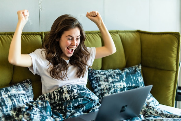 Feliz mujer morena con gesto de victoria usando laptop mientras está sentado en la cama por la mañana