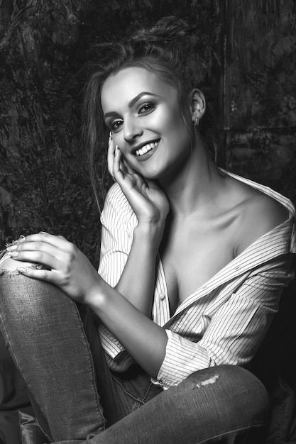 Feliz mujer morena con maquillaje natural viste camisa y jeans posando en el estudio. Retrato en blanco y negro