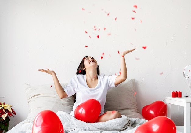 Feliz mujer morena sentada en la cama con globos en forma de corazón rojo lanzando confeti en el aire