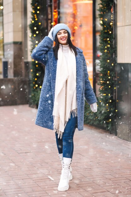 Feliz mujer morena viste gorro de punto rosa claro y bufanda caminando en la ciudad durante las nevadas. Espacio para texto