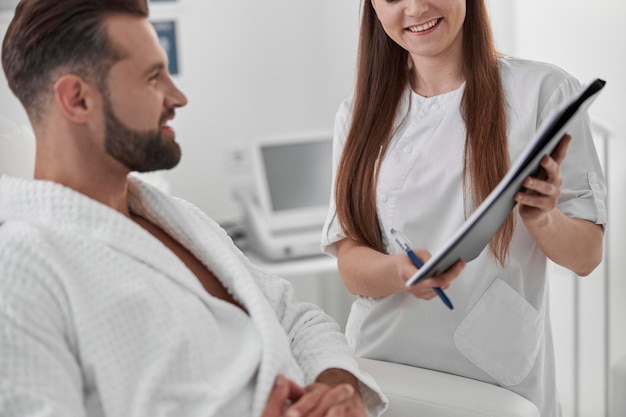 Feliz mujer muestra papel al cliente masculino sentado en un cómodo sillón en la clínica cosmetológica