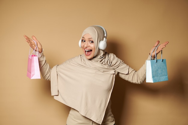 Feliz mujer musulmana levantando los brazos con bolsas de colores en sus manos, sonríe mirando a la cámara