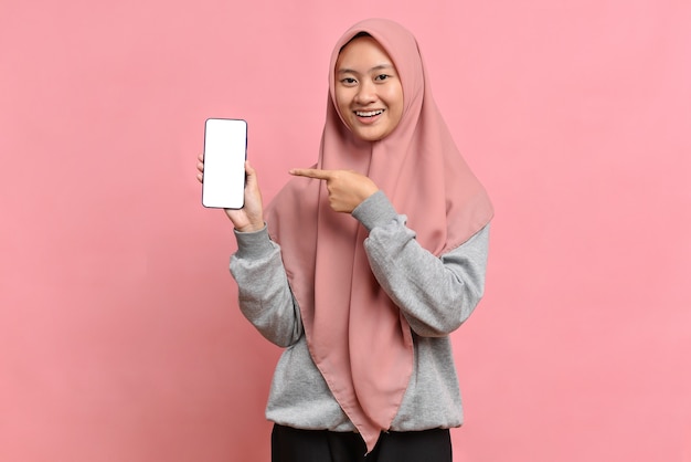 Feliz mujer musulmana señala con el dedo índice en la pantalla en blanco, muestra un dispositivo moderno, vestido con hijab, aislado sobre fondo rosa. Concepto de tecnología