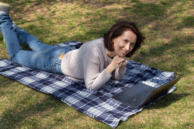 Feliz mujer de negocios acostada sobre una manta al aire libre en el parque trabajando en un fondo natural portátil