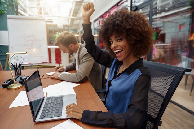 Feliz mujer de negocios africana sentada en su lugar de trabajo en una oficina moderna con antecedentes de colegas