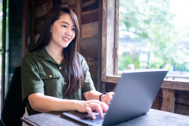 Feliz de una mujer de negocios asiática independiente que usa un vestido verde pardusco con estilo hipster informal que trabaja con una computadora portátil con una taza de café al lado de la ventana de un fondo de casa de madera
