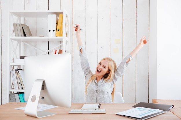 Feliz mujer de negocios emocionada celebrando el éxito mientras está sentada en su lugar de trabajo con las manos levantadas