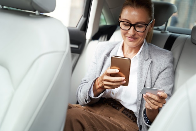 Feliz mujer de negocios exitosa usando anteojos usando su teléfono inteligente y tarjeta de crédito para comprar