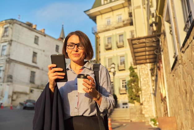 Feliz mujer de negocios madura en vasos con taza de café con teléfono inteligente en la calle de la ciudad Gente de mediana edad de estilo de vida empresarial