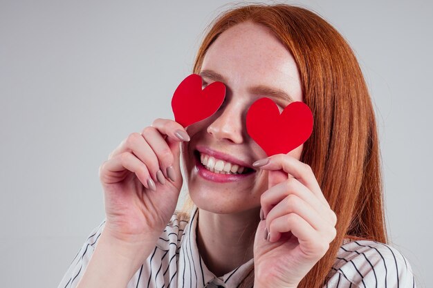 Feliz mujer de negocios pelirroja en una camisa a rayas modelo cierra los ojos con corazones decorativos regalo de San Valentín en el estudio de fondo blanco