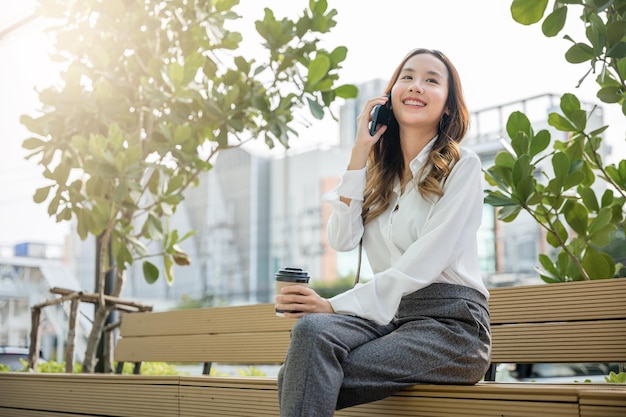 Feliz mujer de negocios sonriente sentada hablando por teléfono móvil fuera de la ciudad de la calle cerca de la oficina, mujer joven de negocios asiáticos usando un teléfono inteligente móvil hablando por teléfono celular al aire libre del edificio corporativo exterior