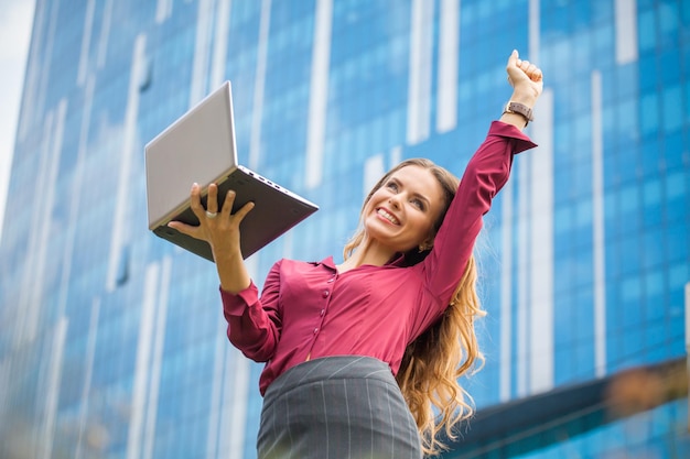 Feliz mujer de negocios sosteniendo una computadora portátil y riéndose en el centro de la ciudad. Mujer celebrando el éxito de su empresa cerca del edificio de oficinas.