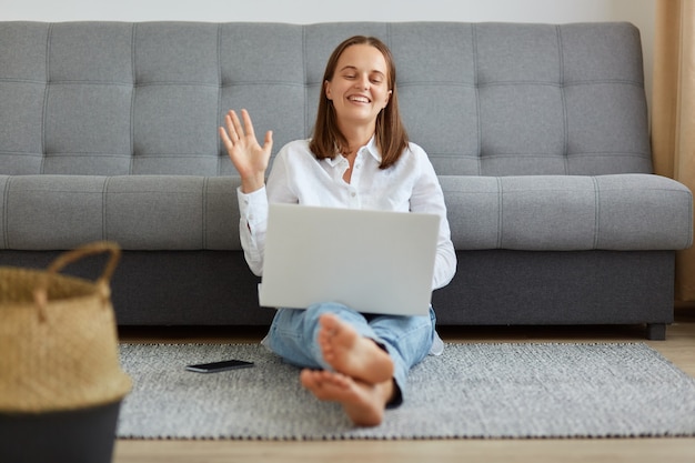 Feliz mujer optimista con camisa blanca y jeans sentado cerca del sofá en la sala de estar, mirando la cámara web de la computadora portátil y saludando con la mano, diciendo hola o adiós, teniendo una videollamada.