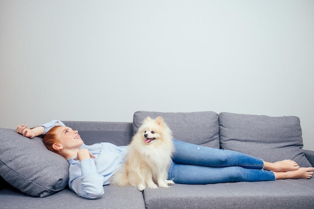 Feliz mujer pelirroja de jengibre con camisa y jeans acostada con su spitz esponjoso blanco descansa en un nuevo sofá plegable gris copyspaseneed aire acondicionado