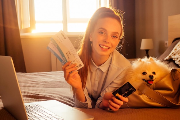 Feliz mujer pelirroja pelirroja acostada en la cama con su lindo perro blanco y esponjoso spitz en una bolsa de viaje que lleva y muestra boletos de vuelo y tarjeta de crédito al atardecer luz de fondo por la noche