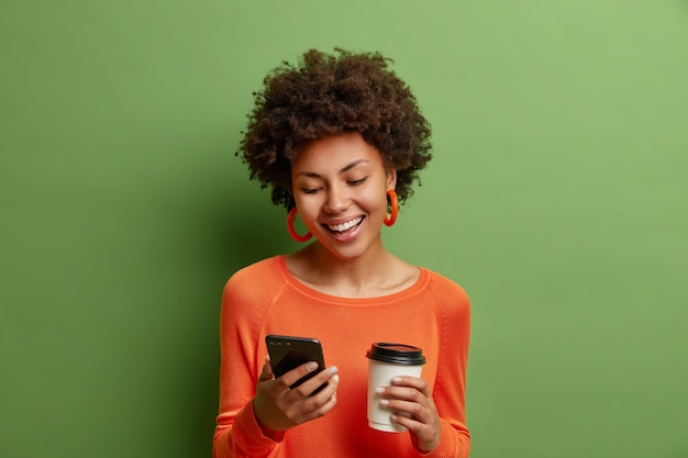 Feliz mujer de pelo rizado encantada sostiene una taza de café de papel y el teléfono inteligente tiene una agradable charla en línea agradable y usa un puente naranja aislado sobre una pared verde