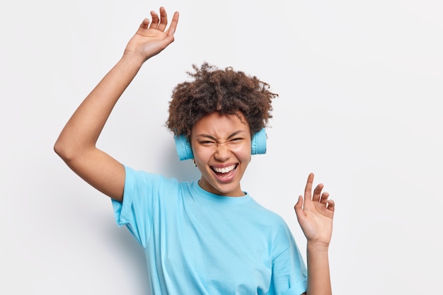 Feliz mujer de pelo rizado vestida con camiseta casual levanta los brazos se divierte disfruta escuchando música a través de auriculares estéreo con buena calidad de sonido expresa emociones positivas aisladas sobre la pared blanca.