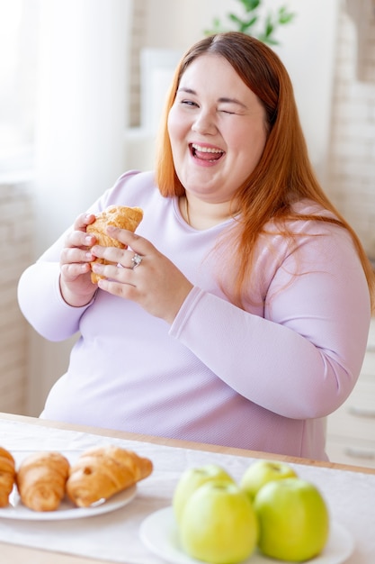 Feliz mujer positiva sentada en la cocina mientras come un delicioso croissant